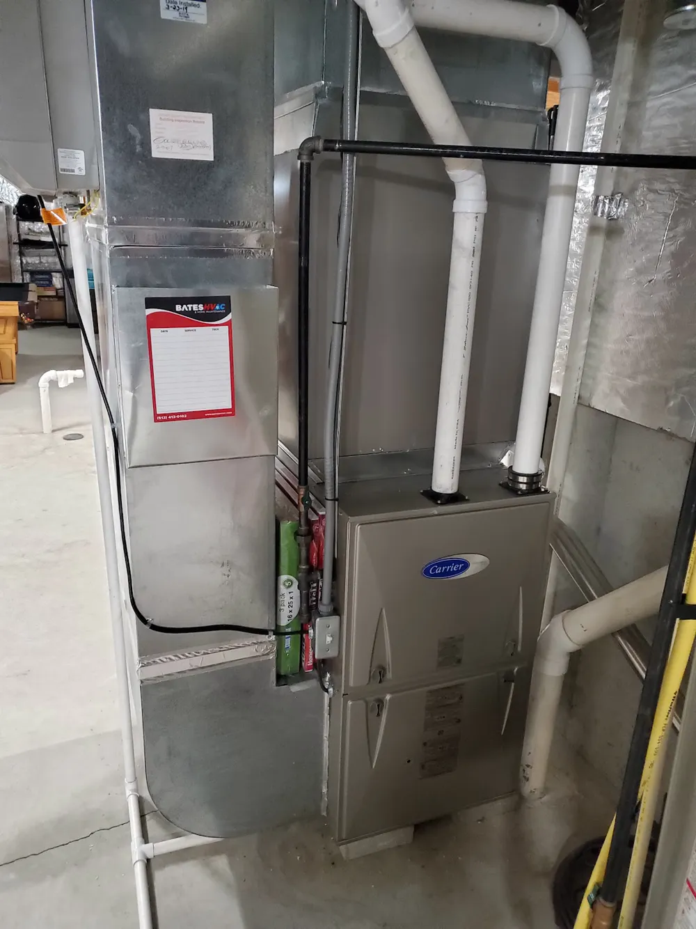 A Carrier gas furnace in a basement with white PVC vent pipes and a Bates HVAC service sticker on the ductwork