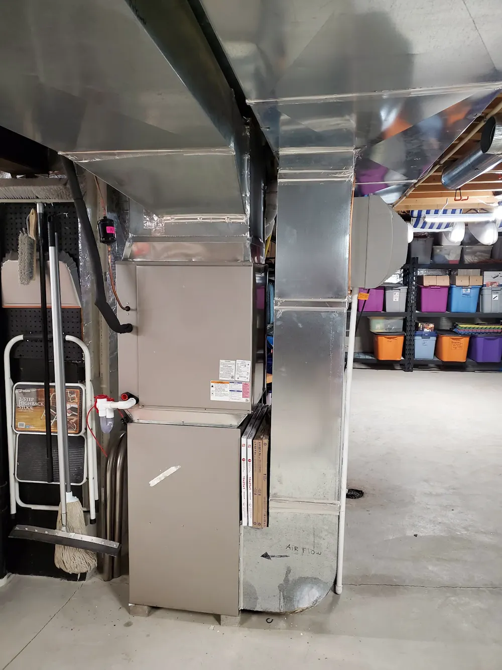 A furnace under a large sheet metal supply plenum and duct trunk in an unfinished basement